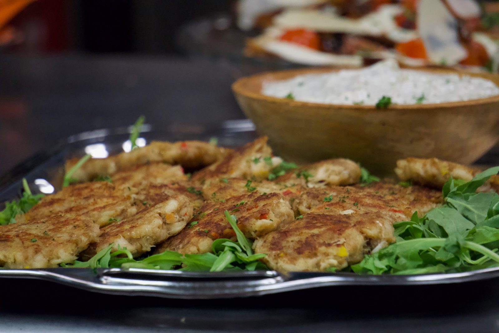 Pan-seared crab cakes arranged on a silver tray over dressed greens, a wooden serving bowl behind them.