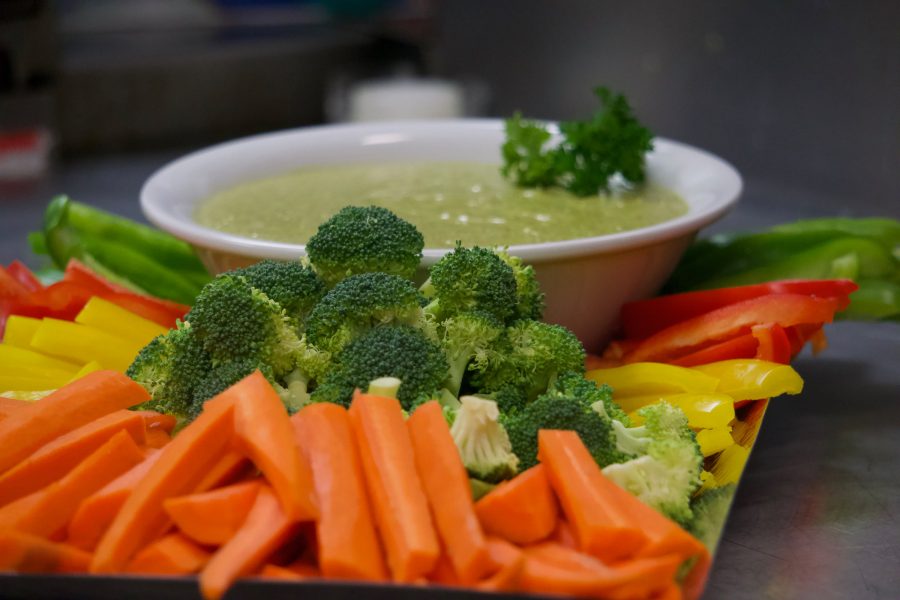 A crudite platter — carrot sticks, broccoli florets and red and yellow pepper strips arranged around a bowl of pale green dip.