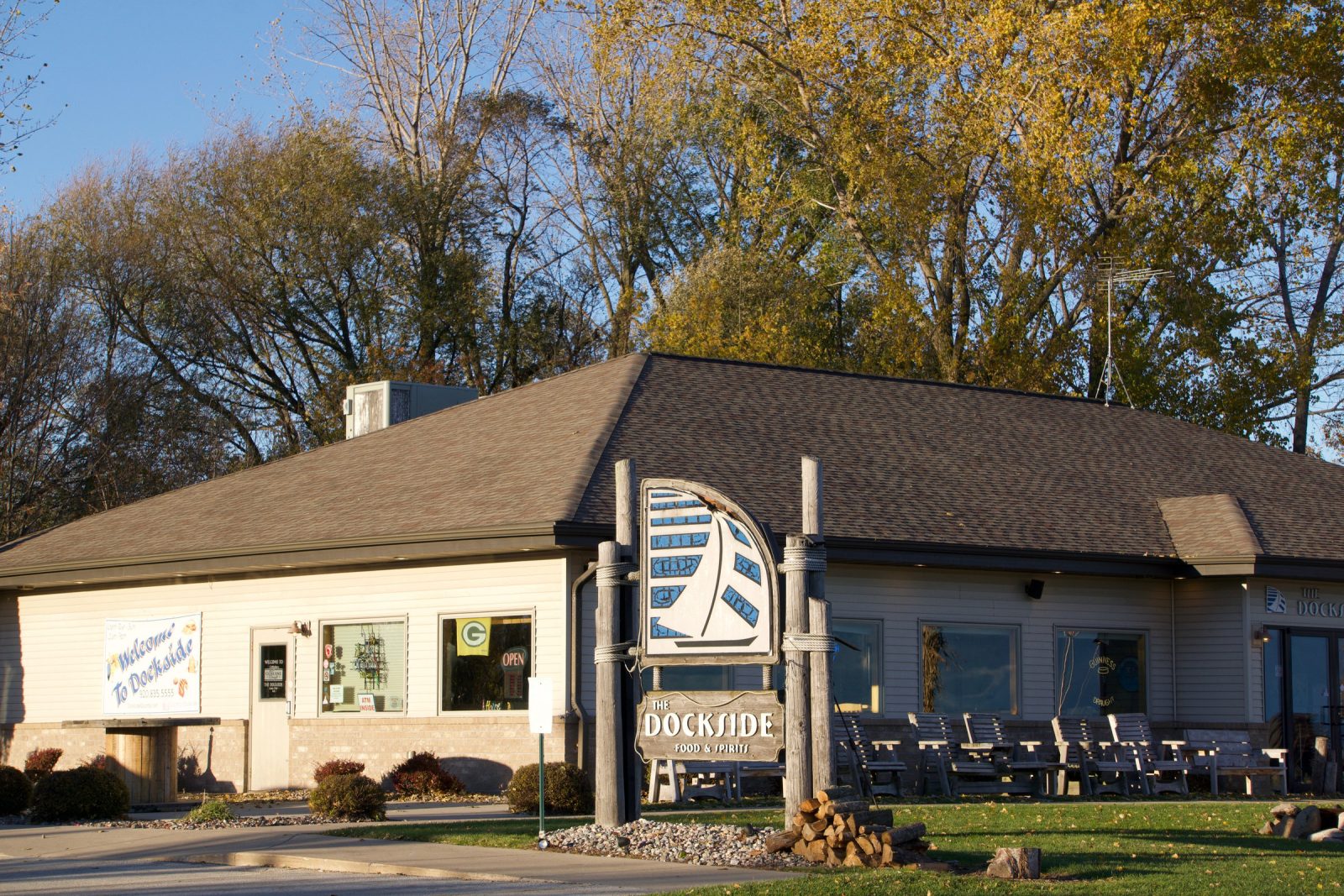 The Dockside from the road — cream siding under a dark roof, a tall sign carrying the restaurant's sailboat badge, patio furniture stacked along the front, bare autumn trees behind.