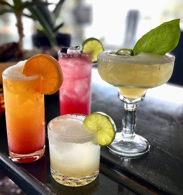 Four cocktails lined up on the bar — a tall orange one with a citrus wheel, a pink one, a coupe garnished with basil and sliced jalapeno, and a lime-garnished rocks glass.