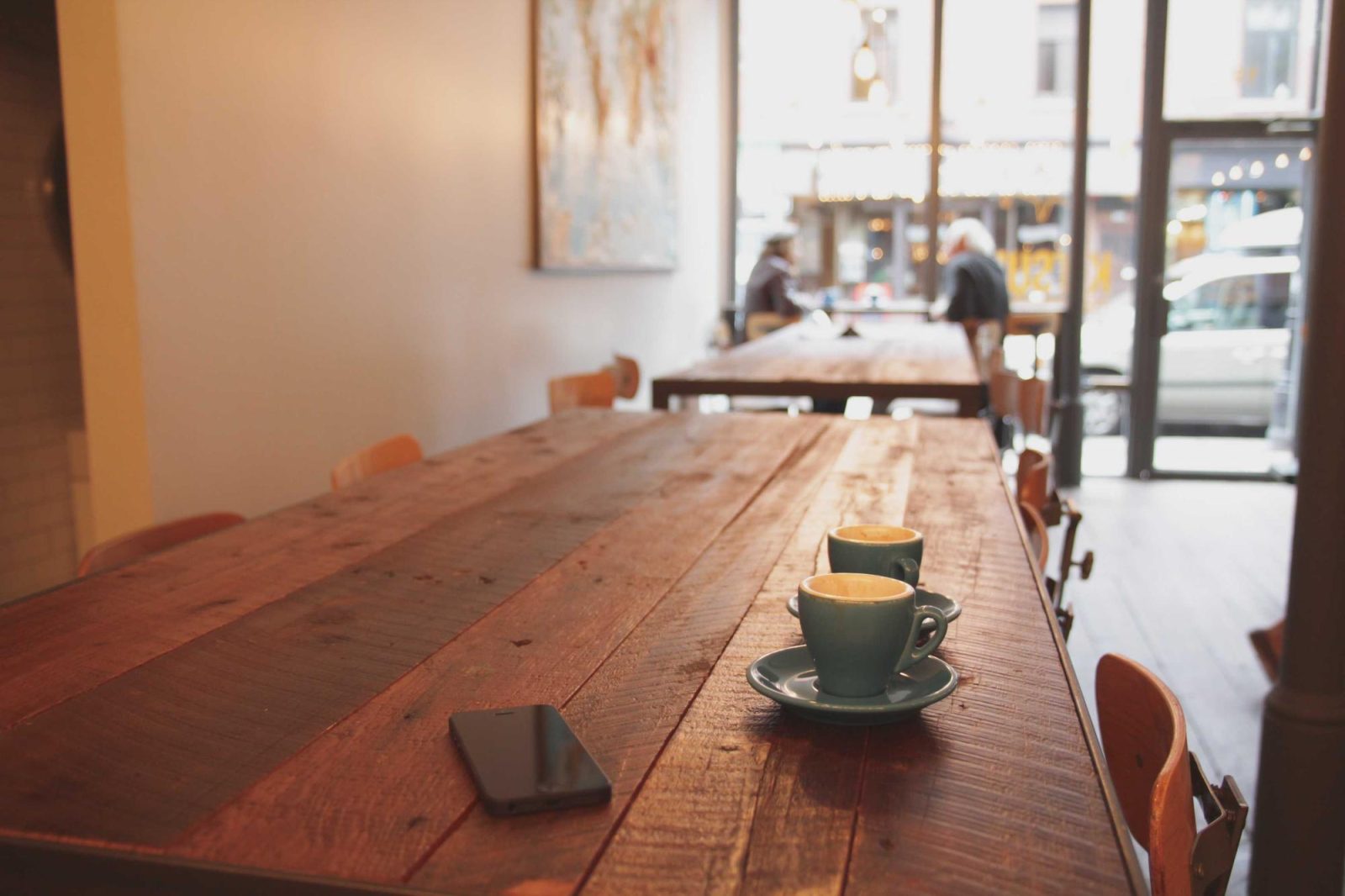 The inside of a restaurant — a long communal wooden table with coffee cups on it, chairs down both sides, daylight from a window at the far end.