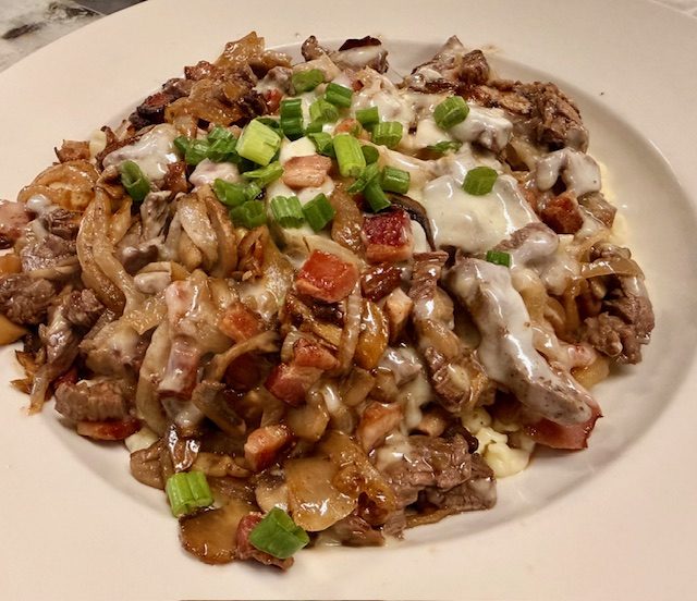 Hand-made spaetzle in a cream sauce with bacon, mushrooms and spring onion, on a white plate.