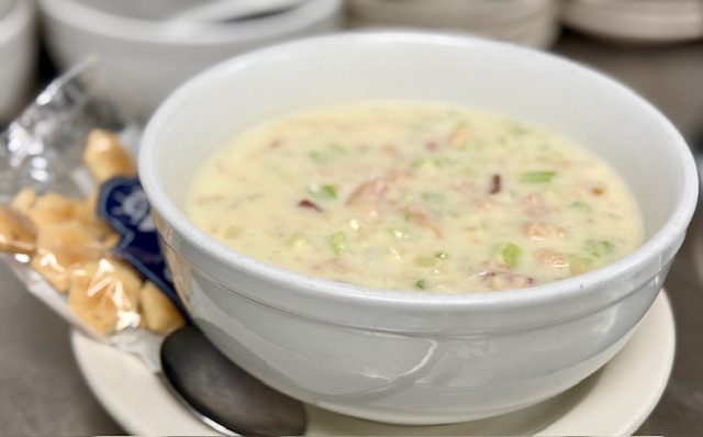 A white bowl of clam chowder scattered with spring onion, a packet of oyster crackers carrying The Dockside's sailboat badge resting beside it.