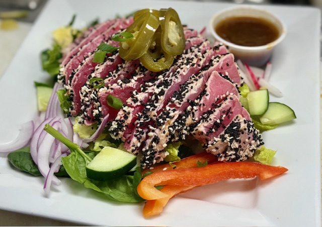 Slices of sesame-crusted seared ahi tuna fanned over mixed greens with pickled jalapenos, red onion and cucumber, a small dish of dressing alongside.