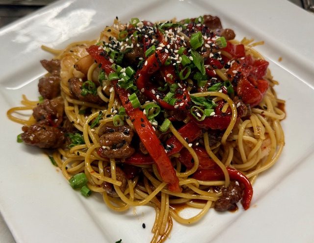 Noodles tossed with sliced beef, red pepper strips, spring onion and sesame seeds, served on a square white plate.