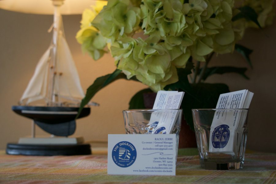 A table by the window — a model sailboat, a vase of hydrangeas, and The Dockside's business cards propped against two rocks glasses printed with the sailboat badge.